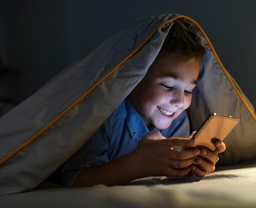 A school-aged boy is using a smartphone with a blanket covering his head.