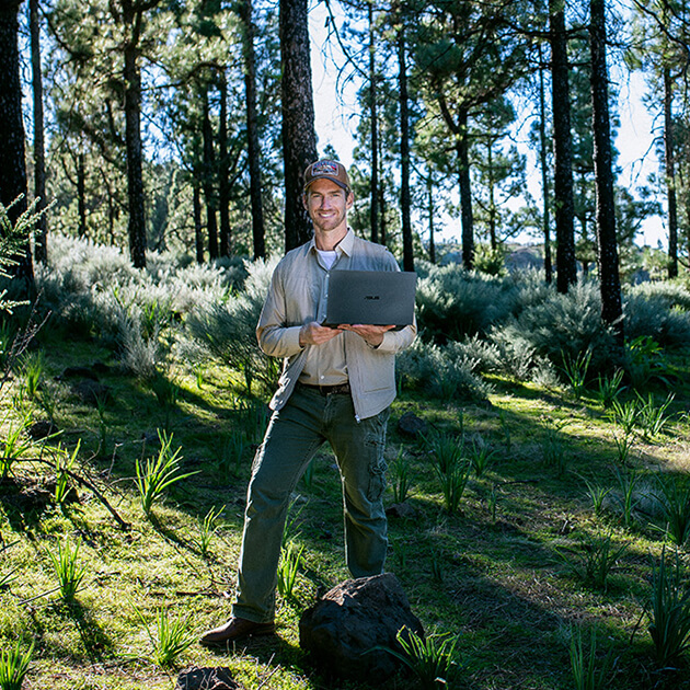 a young, smiling man holding an ASUS laptop while standing in a forest