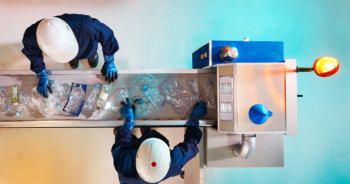 Seen from bird’s-eye view, two men wearing white hard hats are processing plastic parts on a conveyer belt.