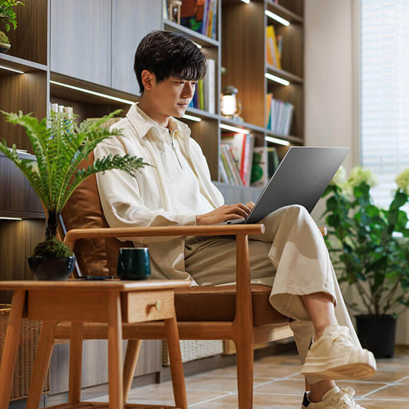 A young man is sitting in a chair using ASUS Vivobook S16 with several plants in the background.