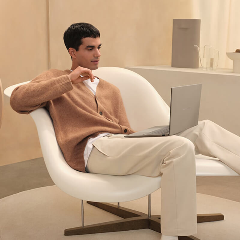 A man sits in a comfortable, laid-back position on a white designer armchair with an ASUS Zenbook A14 laptop on his lap, looking at its screen