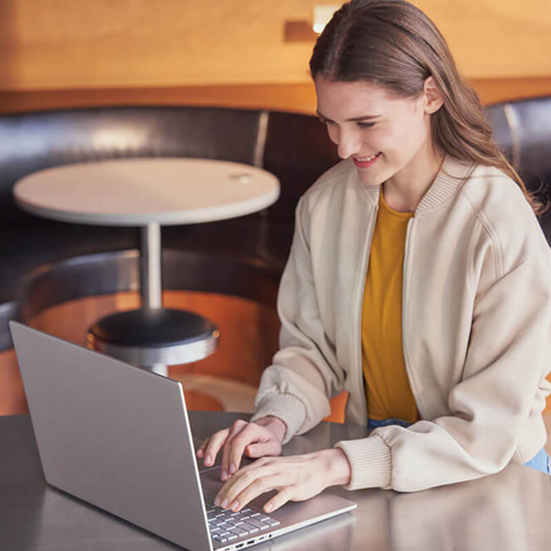 A woman smiles as she sits in a café typing on her ASUS Vivobook S 16 laptop