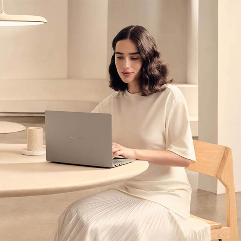 A woman in a beige outfit is sitting at a table using ASUS Zenbook A14.