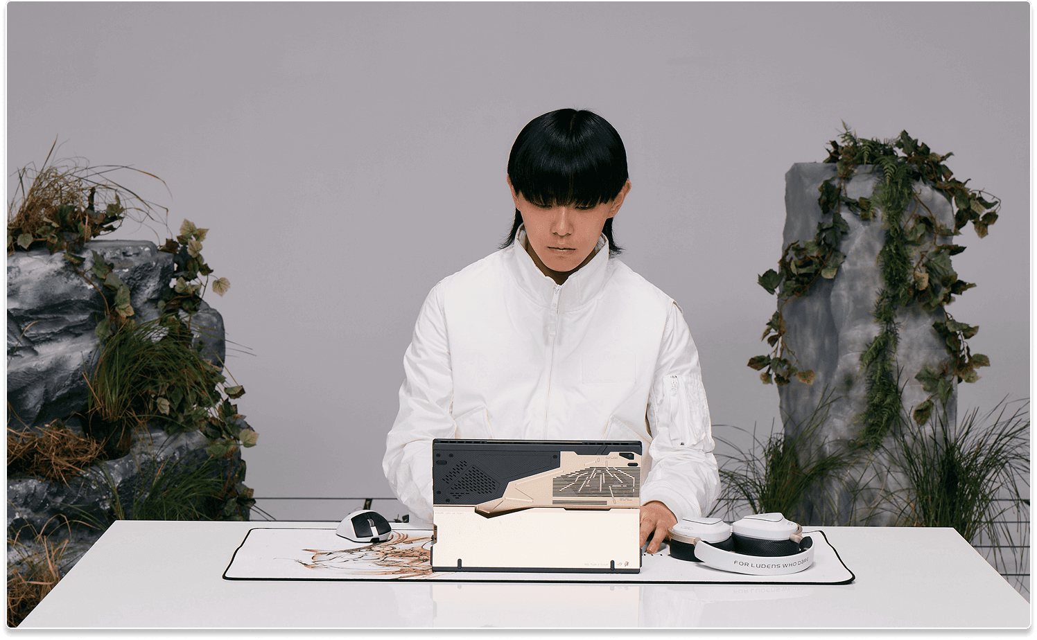 A man in white sitting at a desk typing on the Flow Z13 laptop, which is placed on the Scabbard II XXL KJP mouse pad and next to the Delta II KJP headset and a Keris II Origin KJP mouse