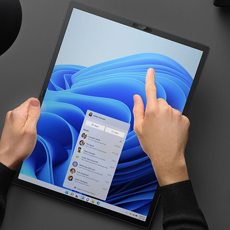 A person using a single hand to hold and a finger to control the touchscreen of the Zenbook 17 Fold OLED with Windows 11 fully extended in Tablet Mode on the desk with elegant black lamp, headphones, watch, clock, pen, cup of coffee and a wallet.