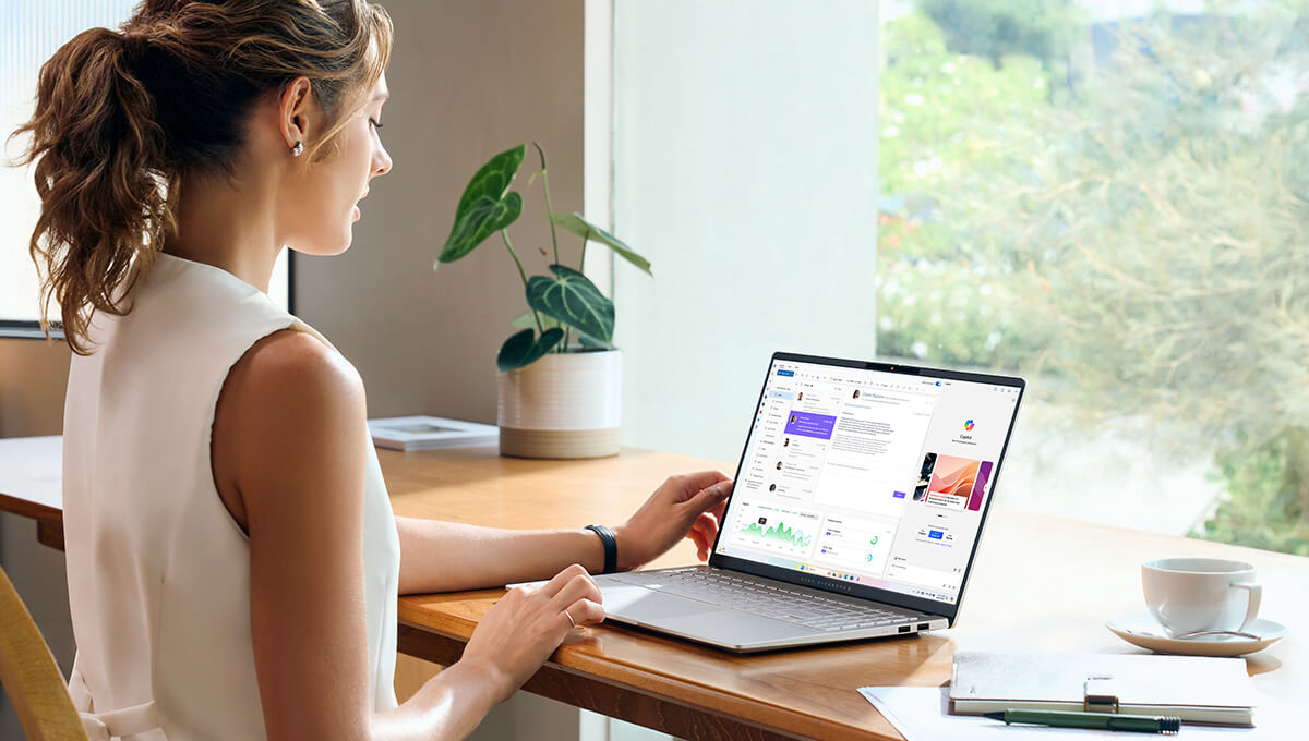 a young woman sitting at a desk near a window with her ASUS Vivobook S 15 laptop while using Copilot AI tool