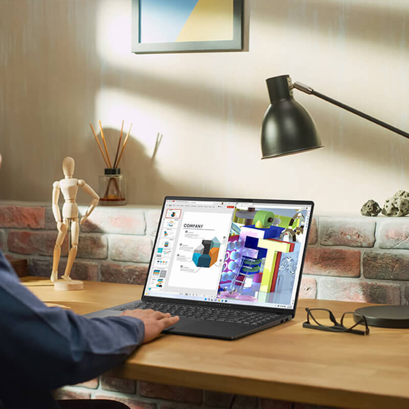 A man in a blue shirt is using an ASUS laptop on a wooden desk with one hand on the trackpad.