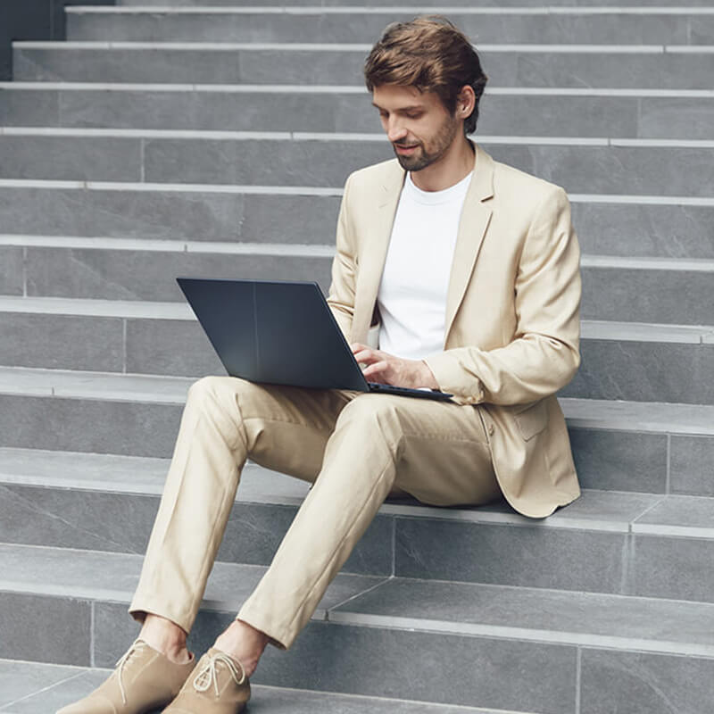 A man in a beige suit is using ASUS Zenbook 14 OLED on his lap while sitting on grey stone stairs.