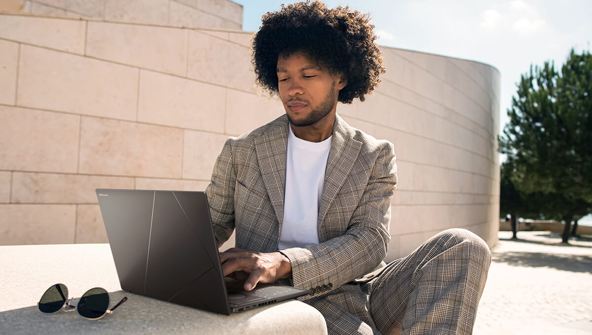 A man in a gray suit is sitting by a stone table outdoors using ASUS Zenbook DUO.