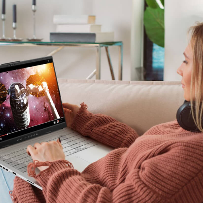 A young woman sitting on a pink sofa with an ASUS laptop on her lap, watching an online video
