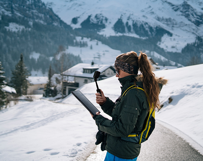 Uma mulher est&aacute; de p&eacute; numa montanha com neve e segura uma c&acirc;mara desportiva e um tablet.