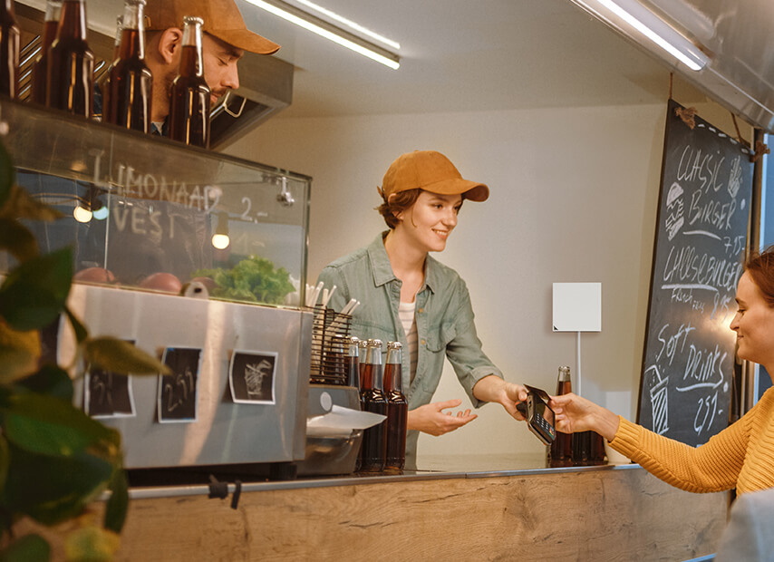Een eigenaar van een foodtruck overhandigt een kaartlezer aan een jonge vrouwelijke klant. De RT-AX57 Go router hangt aan de muur in de foodtruck.