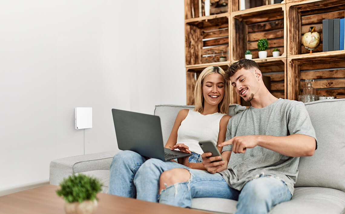 Een lachend jong stel zit thuis op de bank en gebruikt samen een laptop en smartphone.