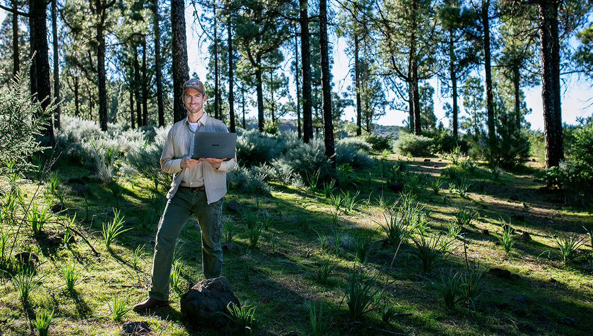 a young, smiling man holding an ASUS laptop while standing in a forest