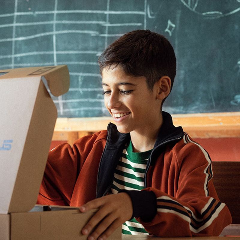 A smiling student opening an ASUS laptop box while sitting in front of a blackboard in a classroom