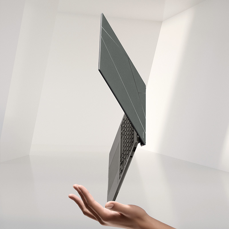 A Zenbook S 13 OLED laptop in Basalt Gray color balancing on its corner on a person’s hand with white walls in the background