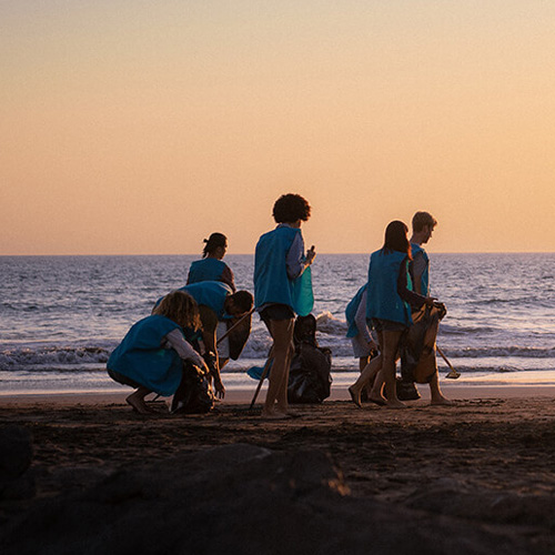 Several people are picking up trash on the beach as the sun sets in the background.