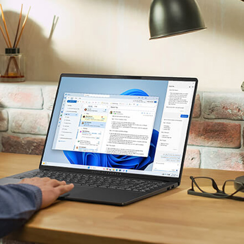 A man in a blue shirt is using Copilot features on an ASUS laptop placed on a wooden desk.